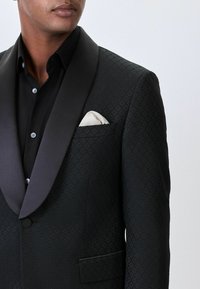 Musthaveid mustaga tekstuuriga teemantmustriga smokkjakk, satiinist tipppäised, ühe nööbiga kinni ja valge taskurätik rinna taskus.