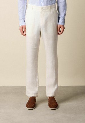 Man draagt crèmekleurige linnen broek, lichtblauw gestreept overhemd en bruine suède instapschoenen, staand op een grijze vloer.