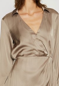 Blusa de satén color arena con un profundo escote en V, mangas largas y un diseño envolvente. Presenta una textura suave y un cuello clásico.