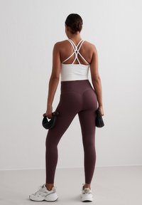 White athletic crop top with crisscross straps, paired with fitted burgundy leggings. The model holds black weights and wears white sneakers.
