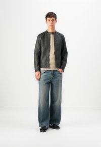 Jeune homme debout portant une veste en cuir noir, un pull beige, un jean bleu à jambes larges et des baskets noires sur un fond blanc uni.
