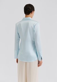 Ανοιχτό μπλε σατινέ πουκάμισο με μακριά μανίκια, γιακά και μανσέτες με κουμπιά. Διαθέτει λεία υφή και καμπυλωτή γραμμή στη βάση.