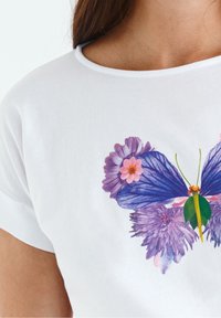 Witte T-shirt met een grote, kleurrijke vlinderafbeelding omringd door paarse bloemen. Zachte stof, ronde halslijn en korte mouwen.