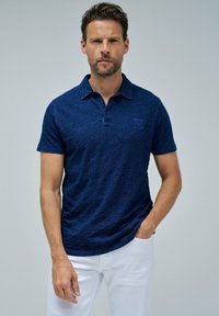 Tummansininen lyhythihainen polo-paita, jossa on teksturoitu kangas, kaulus, kolme nappia ja pieni logo vasemmassa rinnassa.