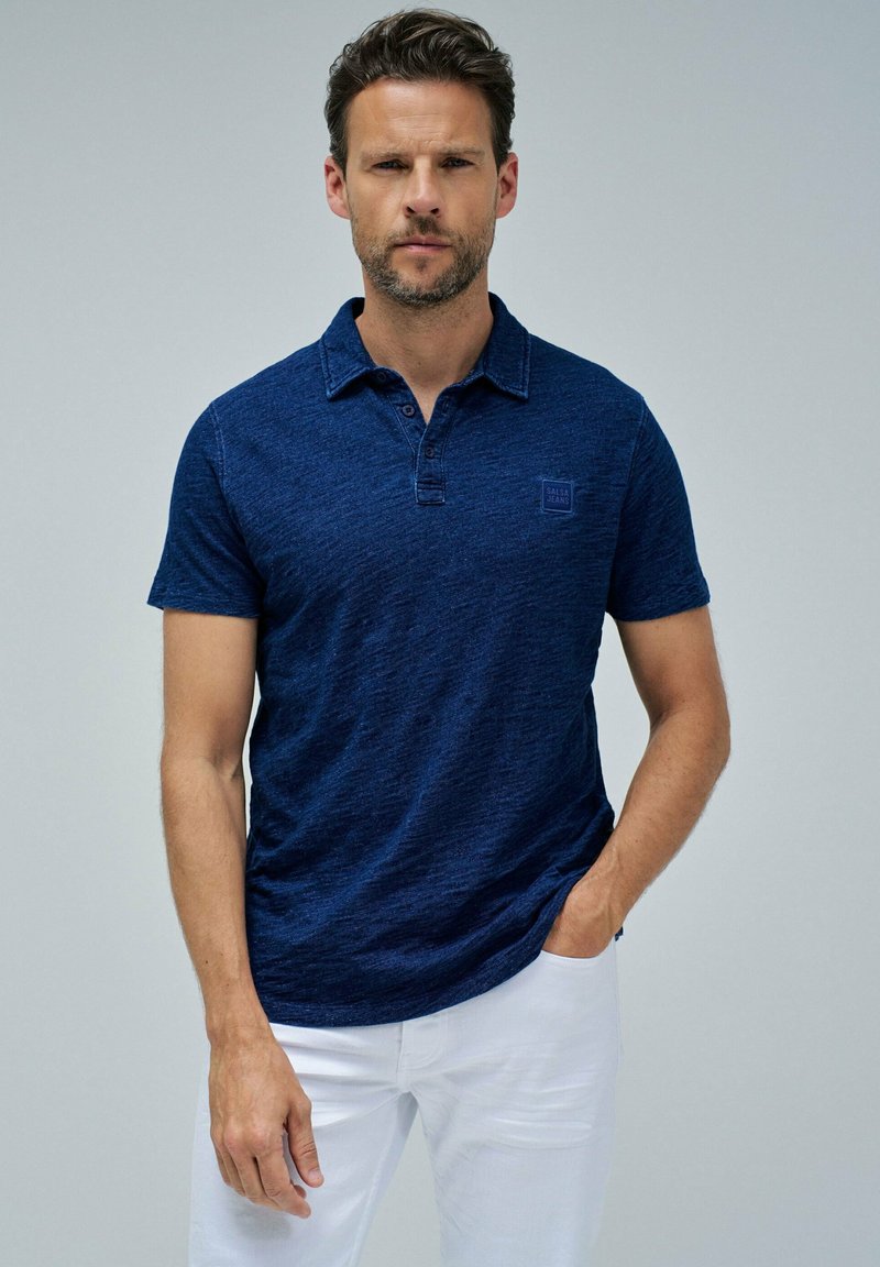 Tummansininen lyhythihainen polo-paita, jossa on teksturoitu kangas, kaulus, kolme nappia ja pieni logo vasemmassa rinnassa.