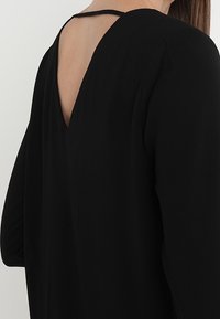 Černý top s dlouhým rukávem a hlubokým výstřihem na zádech, s jemným detailem ramínek. Hladká textura a uvolněný střih zdůrazňují jednoduchost designu.