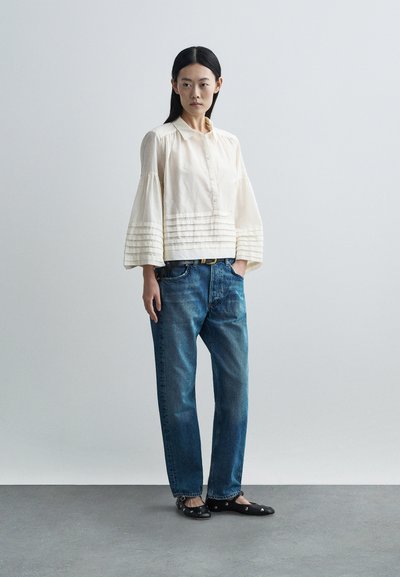 Krémová blúzka na gombíky s nariasenými detailmi na rukávoch, kombinovaná s voľnými modrými džínsami a čiernymi balerínkami s ozdobnými prvkami.