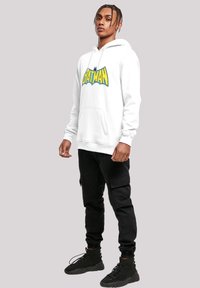 Hvit hettegenser med hette som har en stor blå og gul "BATMAN" grafikk, kombinert med svarte cargo-bukser og svarte joggesko.
