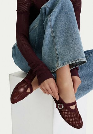 Person in blauen Jeans und burgunderrotem Netz-Oberteil passt passende burgunderrote Netz-Mary-Jane-Schuhe mit silberner Schnalle an, während sie auf einem weißen Block sitzt.