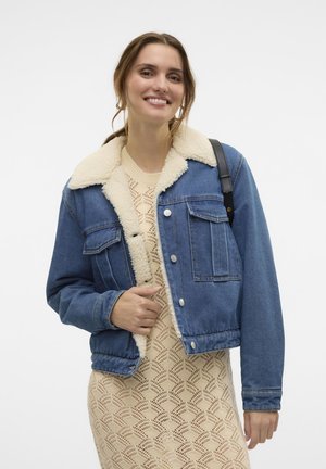 Veste en denim avec un col en fausse fourrure beige, poches avant et fermeture à boutons. Portée par-dessus une robe en tricot crème avec un motif en vagues.