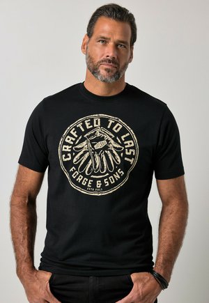 Uomo con capelli corti e scuri e barba, che indossa una maglietta nera con grafica beige di guanti e testo "Crafted to Last Forge & Sons", in piedi con le mani in tasca.