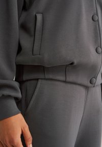 Pelēks ikdienas krekls ar teksturētu audumu, ar noapaļotu apkakli, pogu akcentiem un sānu kabatām, kopā ar atbilstošām sporta biksēm.