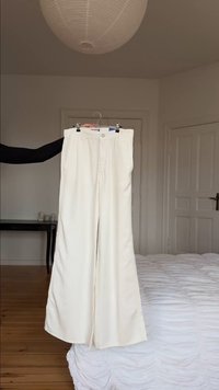 Pantalones anchos blancos colgados en una percha, sostenidos por el brazo de una persona, en un dormitorio con una cama, una mesa y una puerta cerrada al fondo.