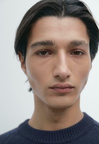 Nahaufnahme eines männlichen Gesichts mit mittlerem Hautton, dunkelbraunen Haaren und braunen Augen. Er hat einen neutralen Gesichtsausdruck und trägt einen dunklen, strukturierten Pullover.