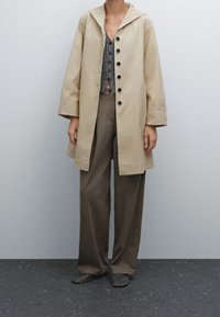 Manteau beige léger arrivant aux genoux avec un grand col et des boutons noirs, superposé sur un haut gris texturé et un pantalon large marron.