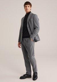 Grau gestreifter Anzug mit einem schwarzen hochgeschlossenen Oberteil. Merkmale sind die taillierte Passform, Revers am Jackett und schwarze Loafers mit Quasten.