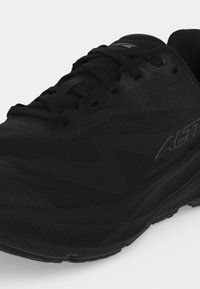 Chaussure de sport noire avec lacets, dessus texturé et semelle épaisse, affichant un marquage latéral en ton foncé sur fond blanc.