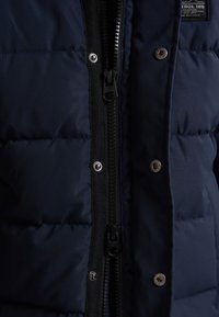 Giacca puffer blu navy con zip frontale e rifiniture nere, bottoni argentati e texture trapuntata. Include un'etichetta del marchio sul petto.