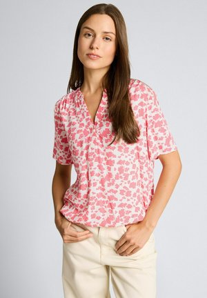 Femme aux longs cheveux bruns portant un chemisier à manches courtes à motifs floraux roses et blancs et un pantalon beige clair, debout les mains dans les poches.