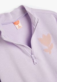 Sudadera de cremallera color lavanda con tela suave. Presenta un diseño floral en rosa en el lado izquierdo del pecho y un tirador de cremallera en contraste.