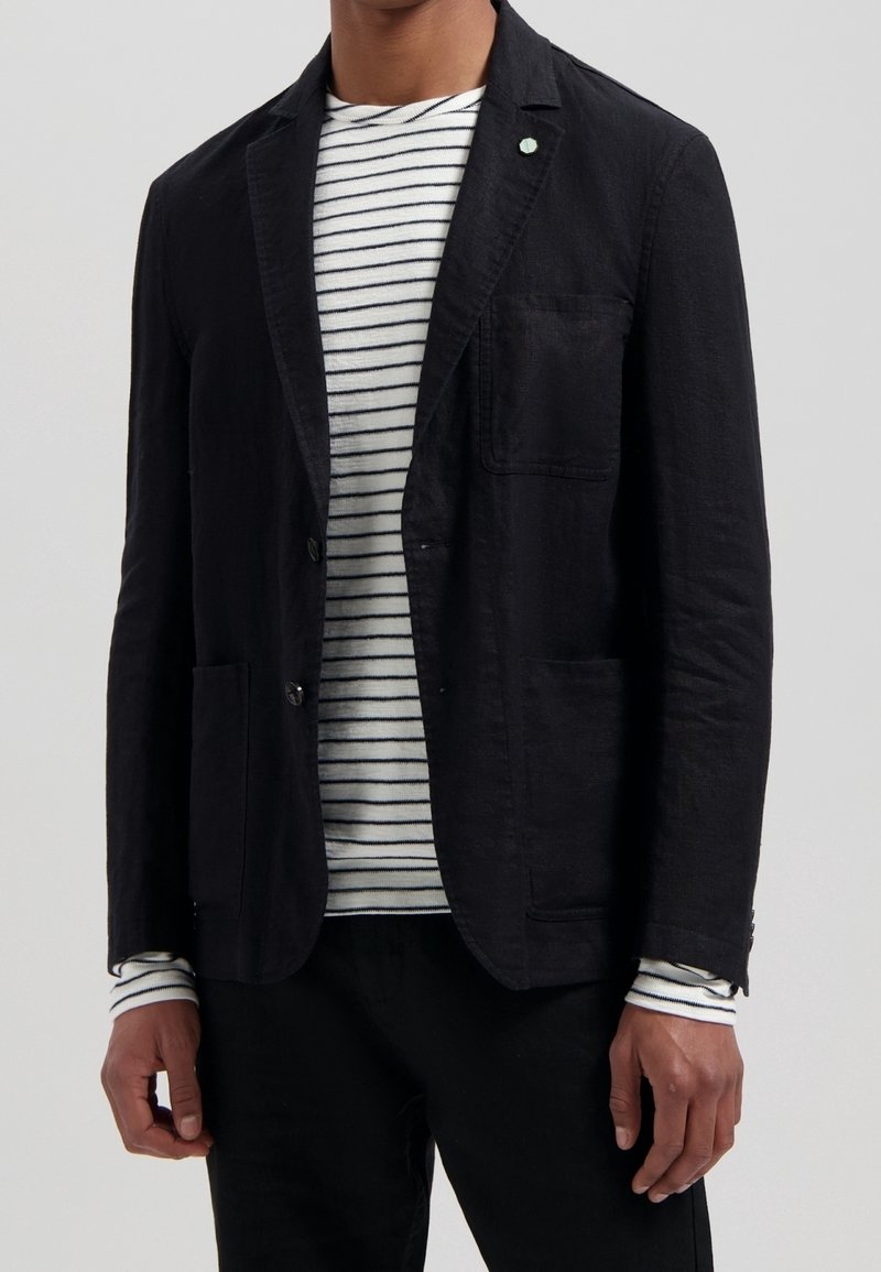 Black linen blazer with a textured finish, single button closure, side pockets, and a small green accent. Worn over a striped long-sleeve shirt.