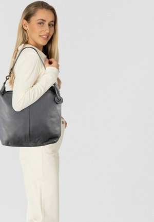 Zwart leren shopper met structuur, voorzien van een enkele schouderband en een ronde logovormige siering, afgebeeld tegen een lichte achtergrond.