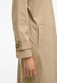 Trench coat beige con taglio dritto, caratterizzato da un polsino con bottoni, tasca laterale e texture liscia.