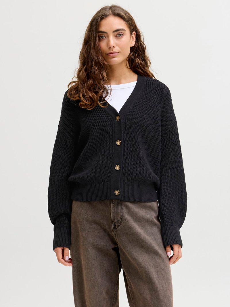 Woman with long curly hair wearing a black buttoned cardigan over a white top and loose brown pants, standing against a plain background.