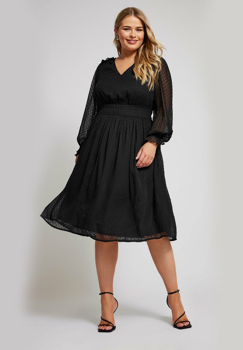 Robe noire avec un décolleté en V, des manches longues transparentes bouffantes et une taille froncée. Le tissu présente un motif à pois subtil.