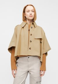 Veste beige courte à manches larges, détails de boutons et col montant, portée sur un pull marron côtelé, associée à un pantalon gris clair.