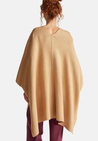 Béžový vlněný pončo s hlubokým výstřihem do V a středovým švem na zádech. Má volný, asymetrický tvar a hladkou texturu.