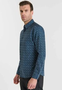 Man in een fitted blauwe langoor-shirt met een klein wit bladerenpatroon en zwarte broek, staand tegen een effen lichte achtergrond.