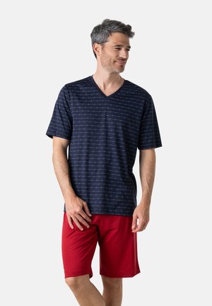Homme portant un t-shirt bleu marine à manches courtes et col en V avec de fines rayures horizontales et un short rouge jusqu'aux genoux, debout et regardant sur le côté.