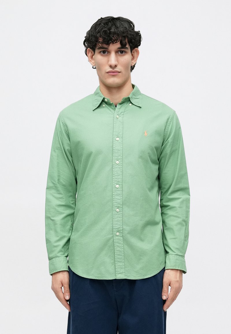 Homem com cabelo preto encaracolado, vestindo uma camisa de mangas compridas verde abotoada e calças azul-escuro, em pé contra um fundo claro e simples.