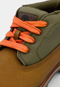 Chaussure en cuir marron avec un dessus en toile vert olive, ornée de lacets orange vif, d'œillets ronds et d'une semelle intérieure texturée.