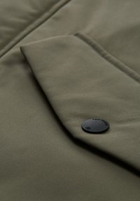 Olivno zelena tkanina s teksturirano površino, s sprednjo žepno zaporko in črnim gumbom z vtisnjenim besedilom.