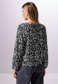 Schwarz-weiße gemusterte Bluse mit langen Ärmeln, rundem Ausschnitt und Raffung auf der Rückseite, kombiniert mit dunkelgrauen Hosen.