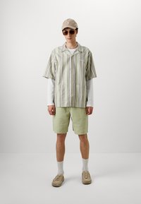 Camisa de manga corta verde y a rayas, pantalones cortos texturizados de color verde claro, gorra beige, gafas de sol redondas y zapatos de lona con estampado.