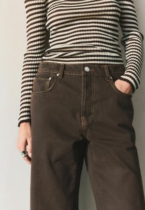 Asmuo, dėvintis rudas aukšto liemens kelnes ir ilgomis rankovėmis megztą marškinėlį su rudomis ir baltomis horizontaliosiomis juostelėmis, ranka kišenėje ir sužadėtuvių žiedu ant piršto.