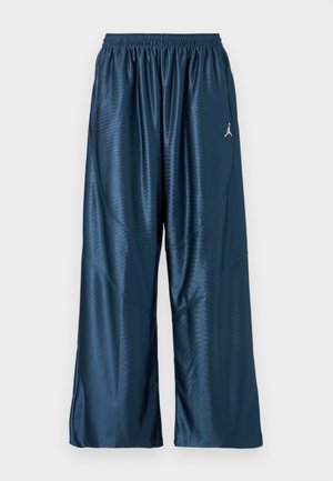 Marineblå sportsbukser med en løs pasform, lavet af skinnende, glat stof. Har en elastisk talje og et hvidt logo på siden.