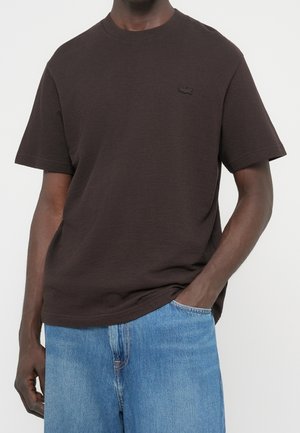 Man draagt een effen donkerbruine korte mouw T-shirt met een klein geborduurd logo en lichtblauwe spijkerbroek, hand in de zak.