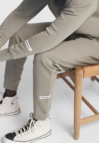 Pantaloni della tuta verde chiaro realizzati in tessuto morbido; presentano polsini aderenti e etichette bianche con testo sulle gambe. Indossati con sneaker alte color avorio.