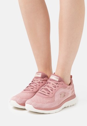 Piernas con zapatos deportivos Skechers rosas con suelas blancas, de pie sobre un fondo blanco.