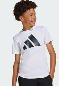 Weißes Baumwoll-T-Shirt mit großem schwarzem Adidas-Logo. Kurze Ärmel und Rundhalsausschnitt, kombiniert mit dunklen Shorts.