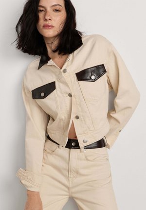 Joven mujer con cabello oscuro lleva chaqueta y pantalones crema con cuello, solapas de bolsillo y cinturón de cuero negro sobre un fondo liso.