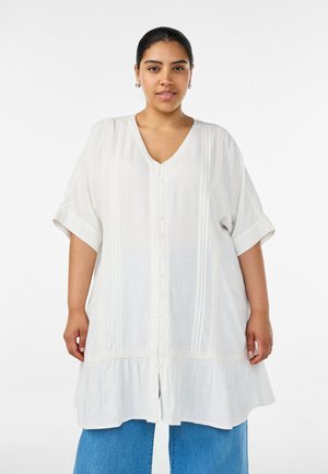 Femme aux cheveux foncés portant une tunique blanche à boutons avec des détails en dentelle et un jean bleu, debout sur un fond blanc uni.