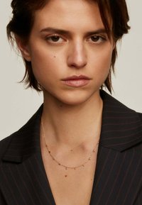 Collar de cadena de oro con pequeños acentos de piedras preciosas coloridas. Llevado sobre un blazer de rayas oscuras, resaltando el escote.