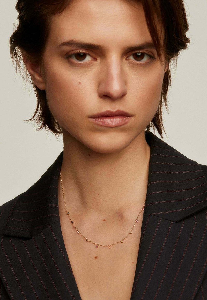 Collar de cadena de oro con pequeños acentos de piedras preciosas coloridas. Llevado sobre un blazer de rayas oscuras, resaltando el escote.