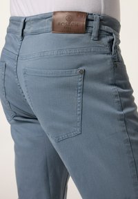 Jeans de denim azul claro con una textura suave, cuentan con una etiqueta de cuero marrón en la cintura, bolsillos traseros y detalles metálicos plateados.