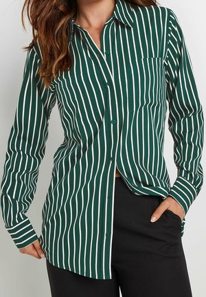 Kobieta w zielono-białej koszuli z pionowymi paskami, zapinanej na guziki, z długimi rękawami i czarnych spodniach, z ręką w kieszeni.